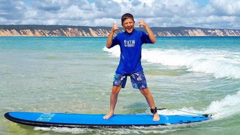 Happy boy giving a thumbs up on a blue surfboard during a Noosa surf lesson in shallow coastal waters, sunny sky in background.