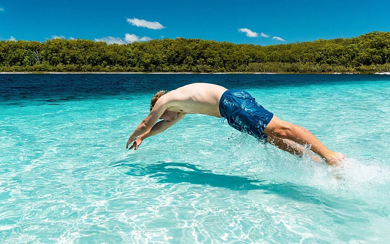 An adventurer in blue swim shorts dives into crystal-clear water by a lush forested shore during a Kgari 3-Day Getaway experience.