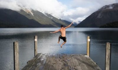 A man dances the Funky Chicken on a wooden jetty before leaping into a serene mountain lake, with scenic peaks and tranquil water.