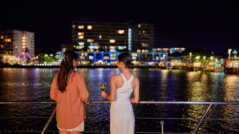 Two women enjoy a night cruise on the Spirit Of Cairns, vibrant harbour lights reflecting as one sips wine onboard the dinner boat.