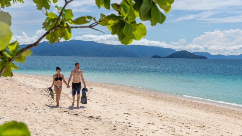 Couple in swimwear walks hand-in-hand on pristine sandy beach with Frankland Island Express Tour kit, sea and mountains in background.