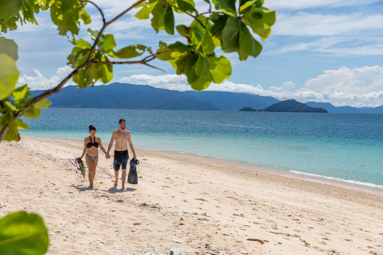 Couple in swimwear walks hand-in-hand on pristine sandy beach with Frankland Island Express Tour kit, sea and mountains in background.