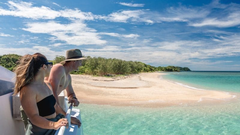 Couple on Frankland Island Express Tour boat gaze at pristine sandy island, surrounded by crystal-clear turquoise water and blue sky.