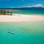 Two adventurers paddleboard in crystal-clear turquoise waters by a pristine sandy island on a Frankland Island Express Tour.