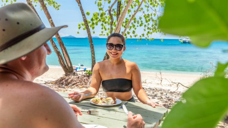 Couple dining at a picnic table on sunlit Frankland Island beach, savouring lunch amid vibrant coastal scenery on Express Tour.
