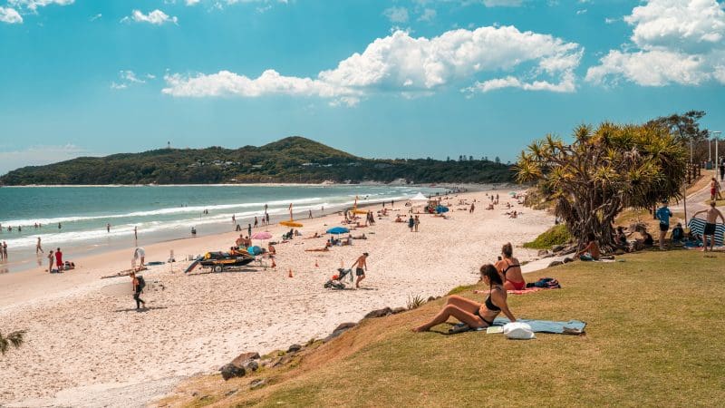 Travellers unwind on a sunlit grassy hill and white sandy beach beside the ocean, along the Sydney to Cairns East Coast adventure tour.