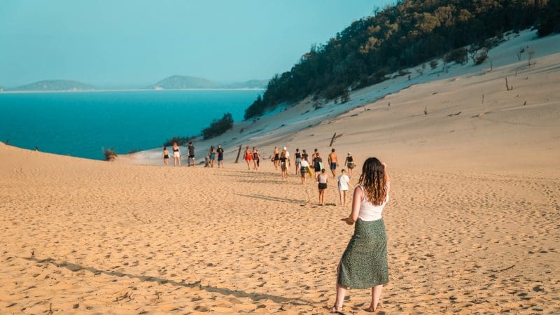 Adventurous woman treks golden sand dunes towards a tour group on the epic Fully Guided Sydney to Cairns East Coast 3-Week adventure.