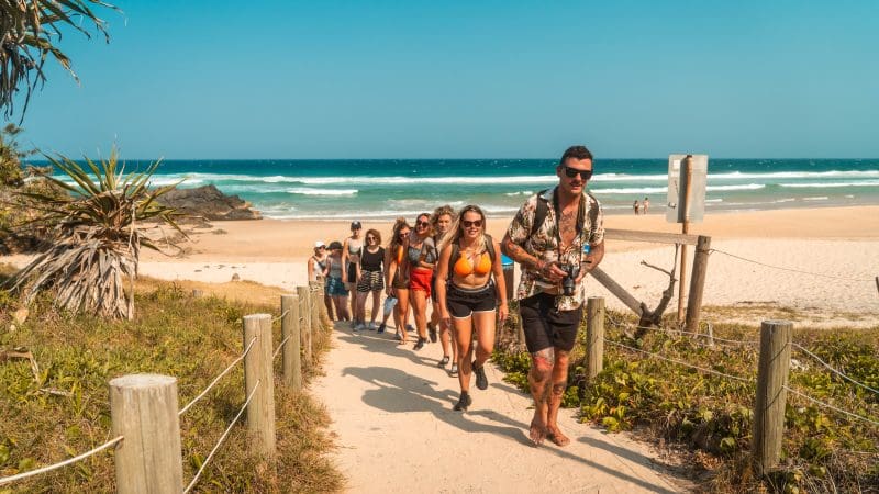 Travellers on the Fully Guided Ultimate East Coast Sydney to Cairns 5 Weeks tour hike a scenic sandy path from the beach under blue skies.