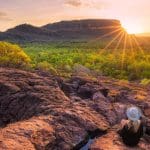 Woman wearing a hat sits on rugged rocks at sunset during a 4-Day Kakadu Katherine Gorge Adventure, enjoying breath-taking views.