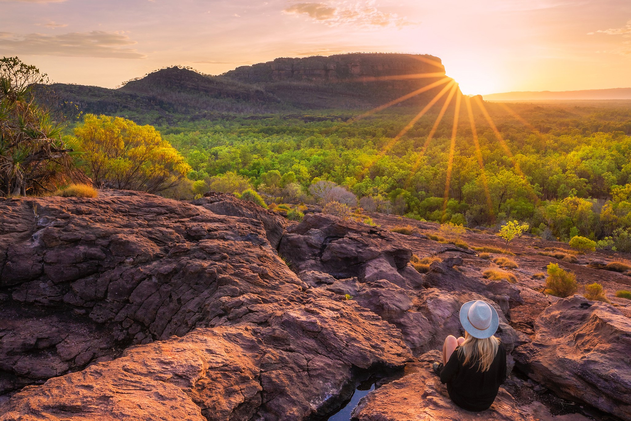 Woman wearing a hat sits on rugged rocks at sunset during a 4-Day Kakadu Katherine Gorge Adventure, enjoying breath-taking views.