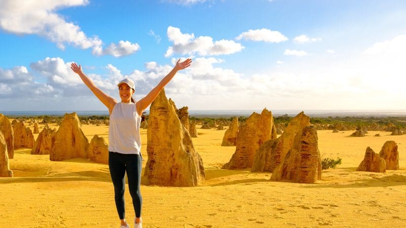 Traveller with arms raised stands amid limestone Pinnacles on a Perth day tour, dramatic clouds overhead, experiencing WA’s unique landscape.