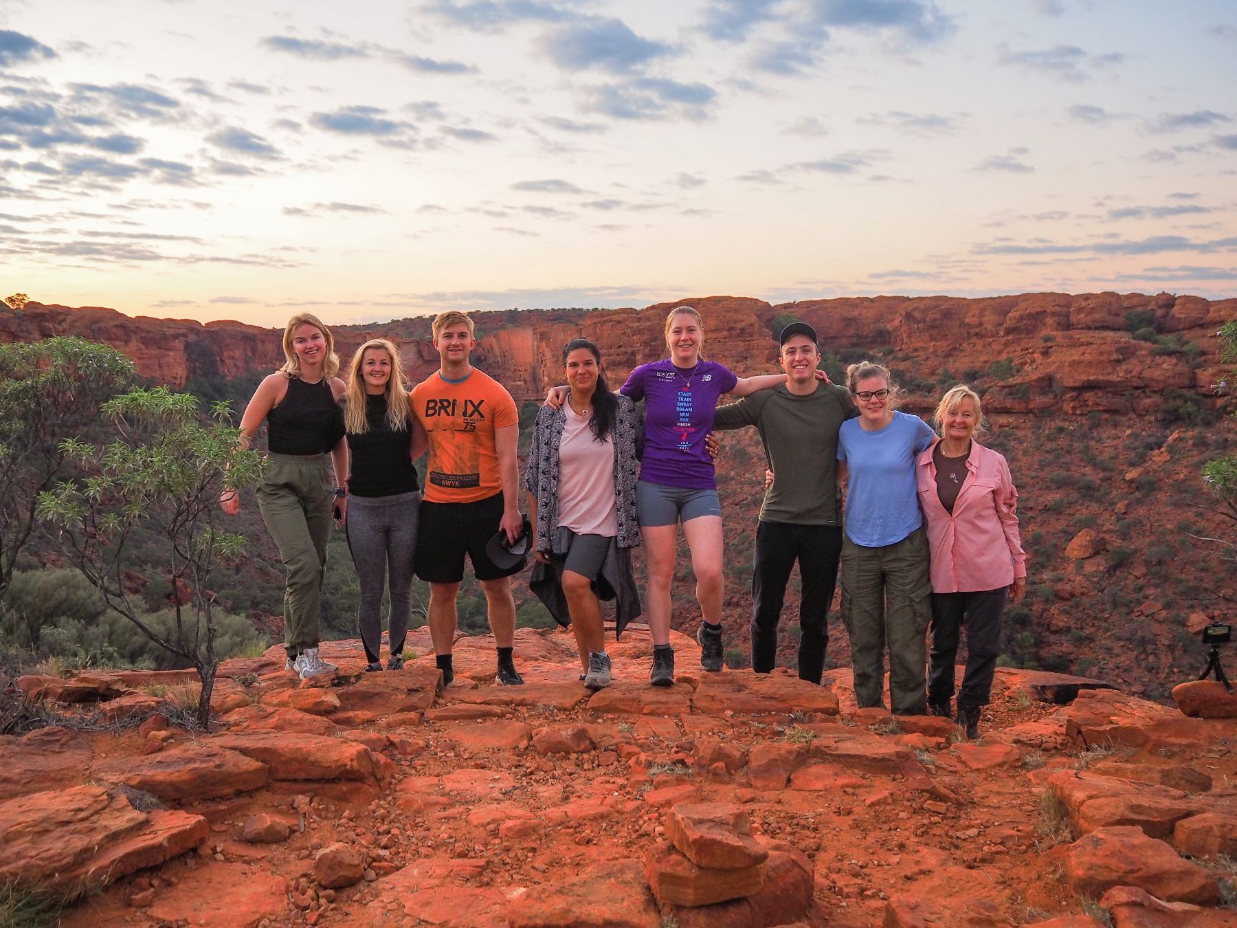 Group of eight adventurers smiling on a rocky sunset cliff during a 7 Day Adelaide to Uluru Untamed Escapes tour experience.