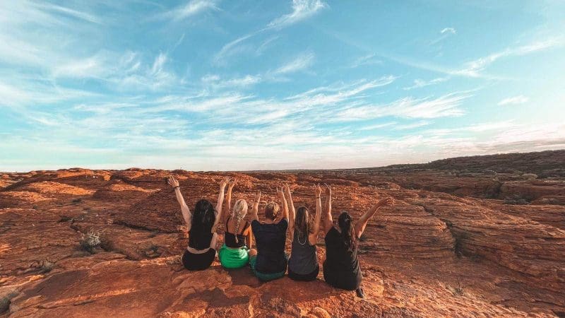 Five travellers relax on vivid red rocks under an expansive sky, enjoying the 7 Day Uluru to Adelaide Experience by Untamed Escapes.
