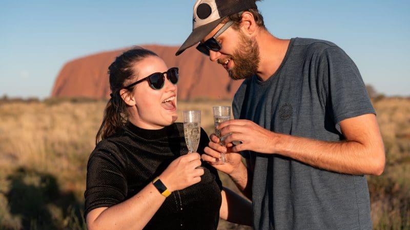 Two travellers in sunglasses toast champagne and smile outdoors on the 7 Day Uluru to Adelaide adventure by Untamed Escapes tour.