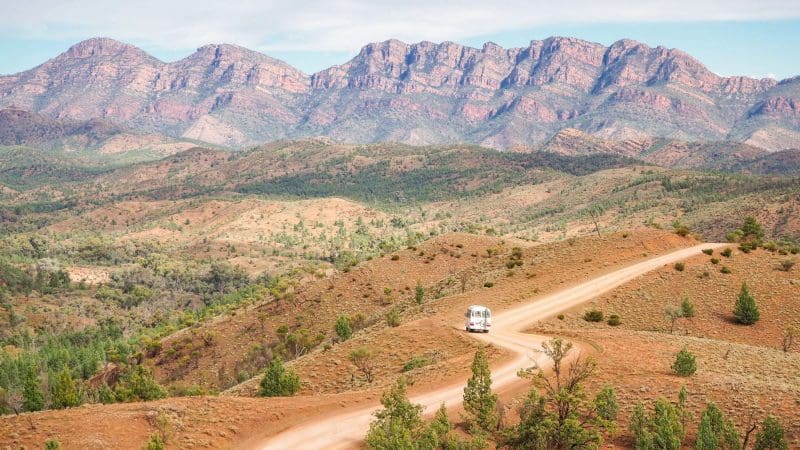 A white campervan travels a scenic dirt road on the 7 Day Uluru to Adelaide tour by Untamed Escapes, showcasing adventure and outback views.