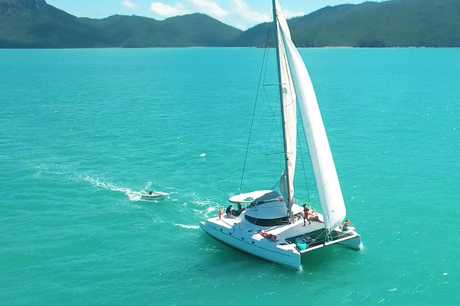 Luxury white catamaran glides on turquoise Whitsunday waters near lush green hills—Entice 2 Day 2 Night Sailing adventure in sun.