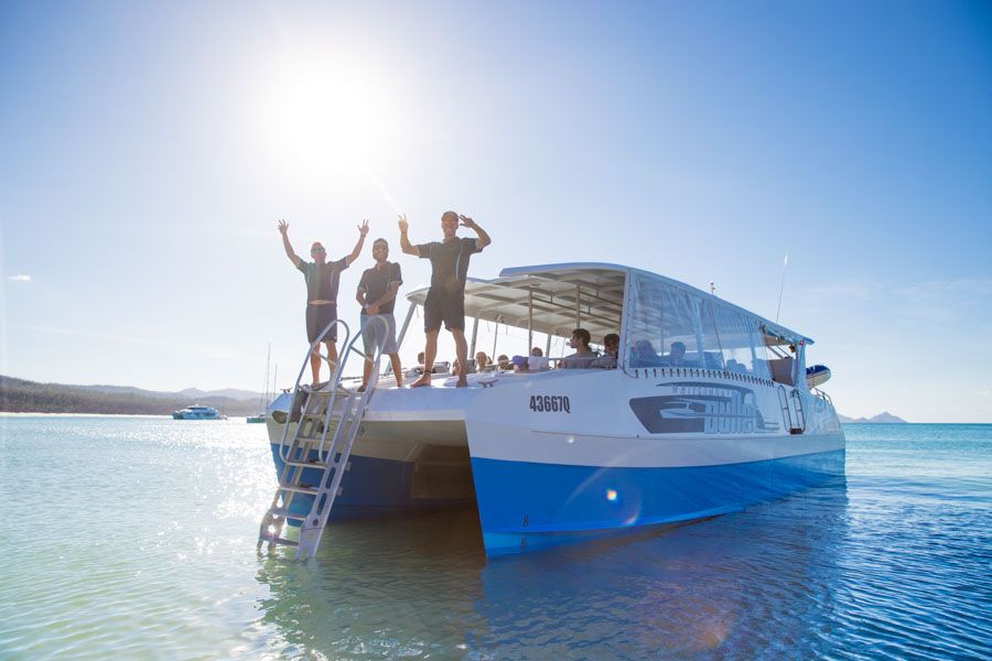 Three smiling travellers wave from the deck of Whitsunday Bullet on a sunny, 1-day Whitsunday Sailing adventure in clear shallow waters.