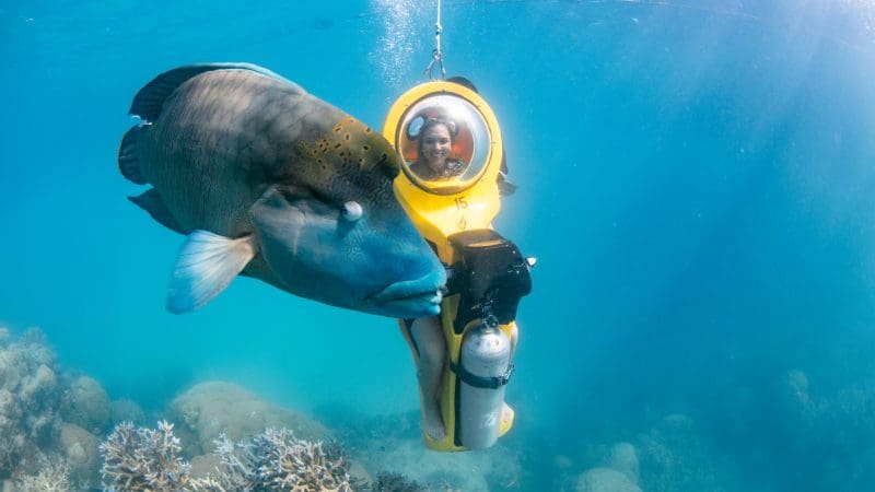 Adventurer explores Great Barrier Reef on a yellow helmet-scooter, surrounded by vibrant coral reefs and a large tropical fish.
