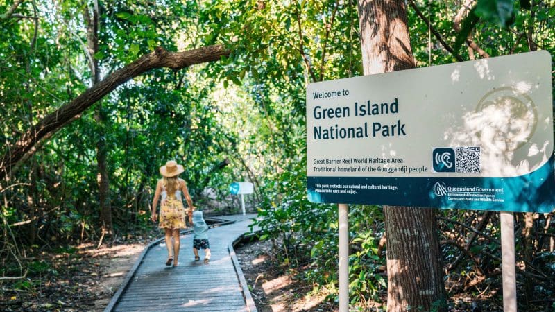 A woman and child stroll along a wooden boardwalk by a Green Island Transfers sign, with lush tropical trees creating a scenic backdrop.