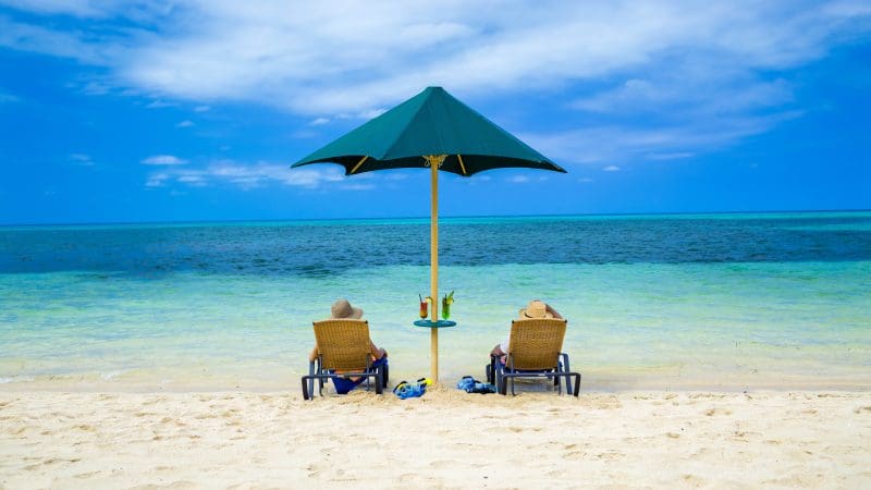 Two people lounge under a green parasol on Green Island, cocktails in hand, ocean views behind them, enjoying their island transfer.