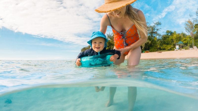 Smiling woman in straw hat assists toddler paddling in crystal-clear Green Island waters, sunny beach backdrop, perfect family day.