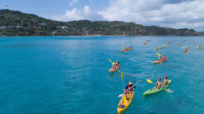 Adventurers kayaking in crystal-clear blue sea waters on a guided tour, passing stunning coastal hills and picturesque seaside houses.