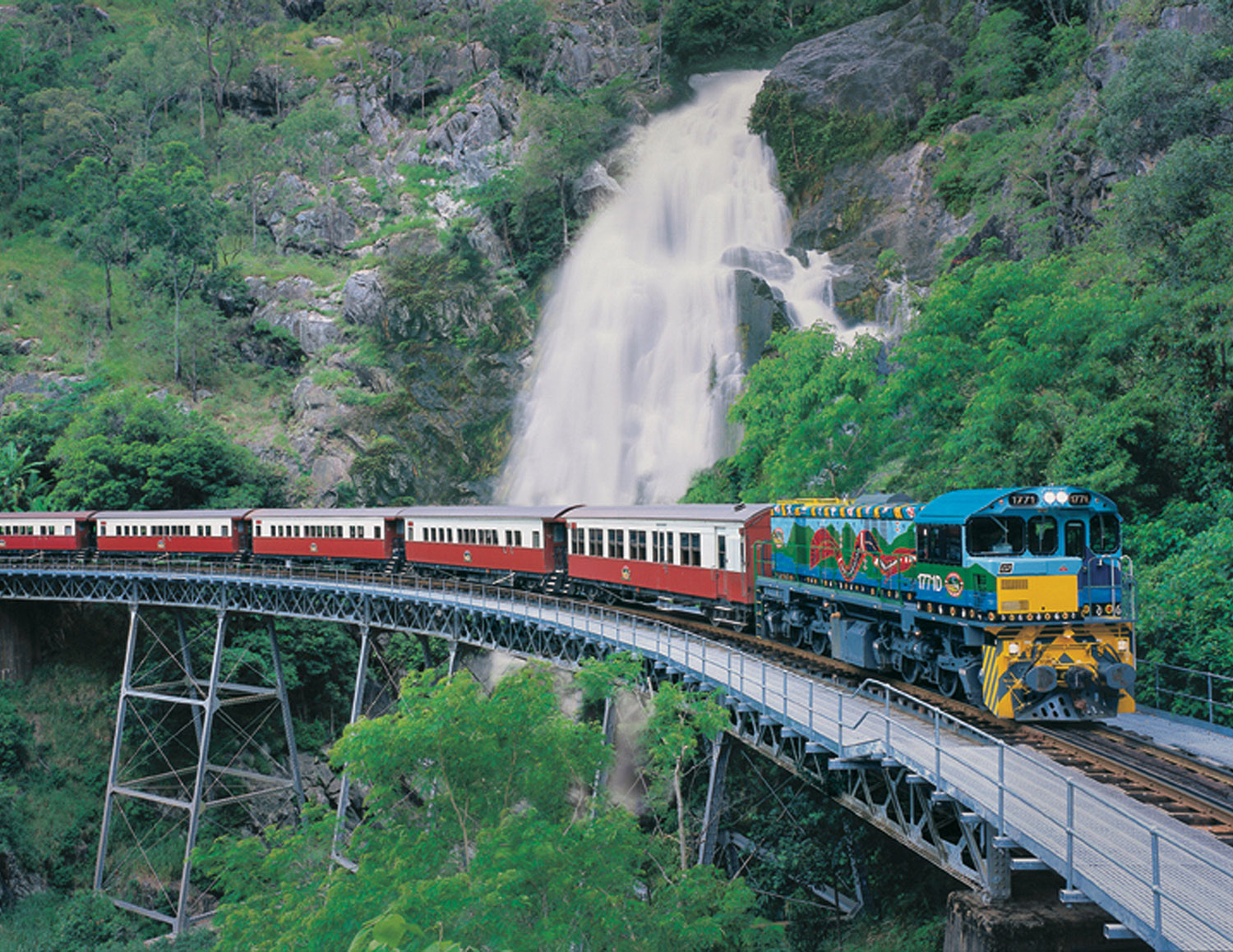 Kuranda Scenic Railway’s colourful Kuranda Deluxe train crosses a bridge near a waterfall and vibrant, lush green rocky hills in Australia.