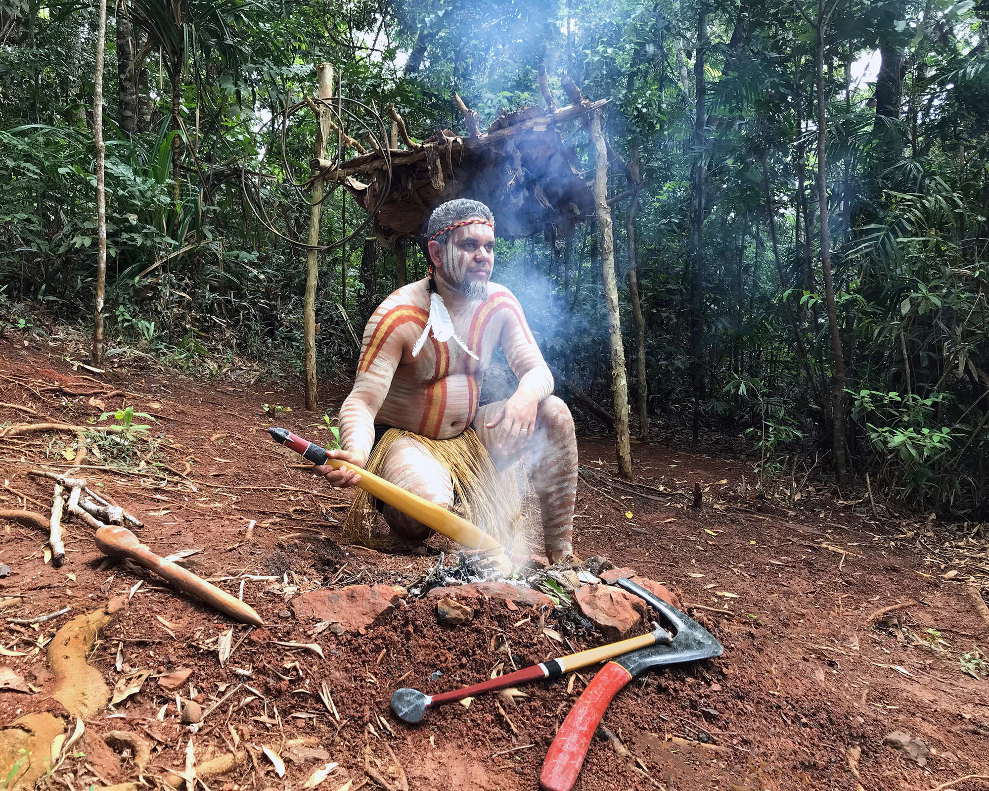 An Indigenous man in vibrant traditional attire sits by a fire, as featured on Express Kuranda Later Departure cultural tour experiences.