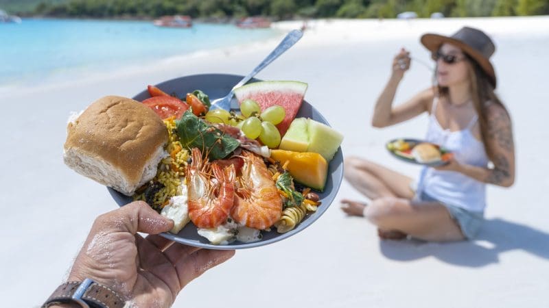 Gourmet seafood, fresh fruit, and artisan bread on a plate at Whitehaven Beach; woman enjoys meal by the shore after snorkelling.