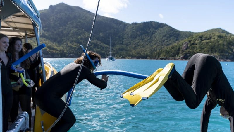 Group in wetsuits on Big Fury Whitsundays boat; one snorkels in crystal-clear water as others smile and watch, enjoying the adventure.