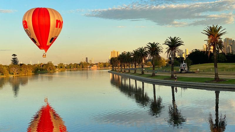 Soar in a red and white hot air balloon above a tranquil Melbourne lake at sunrise—book an unforgettable 1-hour balloon flight.