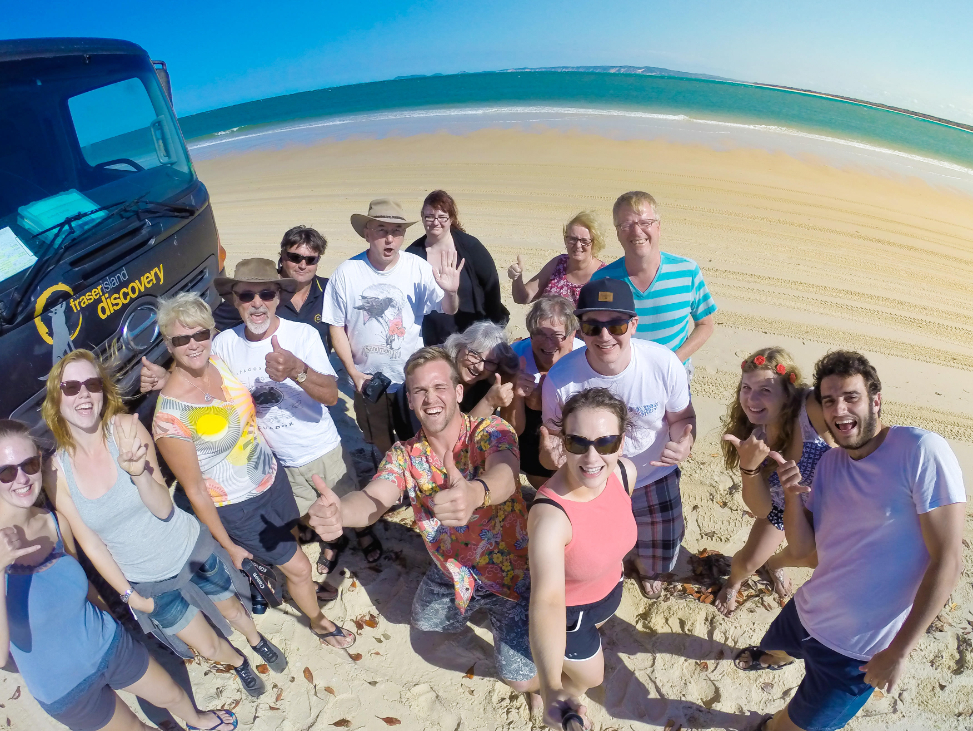Tour group posing and smiling on Rainbow Beach during K'gari Day Tour with a black SUV, clear blue sky, bright sunny weather.