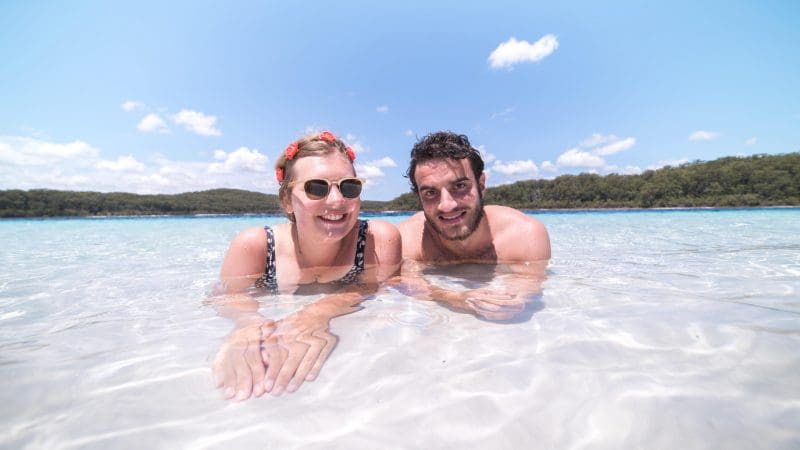 Happy couple wading in crystal-clear, shallow waters on a sunlit beach during Kgari Day Tour from Noosa, perfect for adventure.