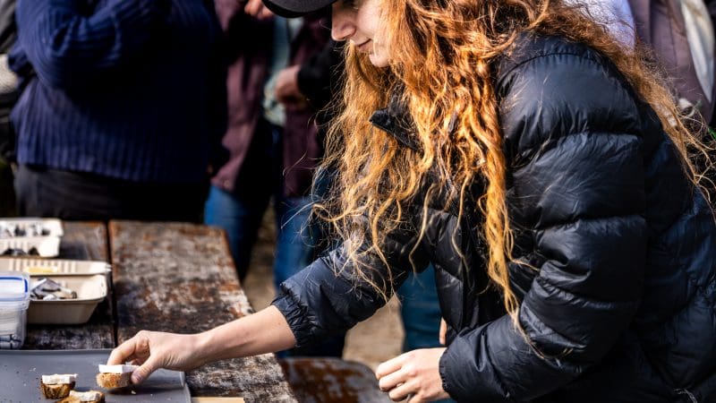 A woman in a black jacket savours gourmet cuisine from a tray at a rustic table during the Bruny Island Food Sightseeing Tour.