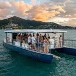Experience a Sundowner Sunset Cruise on a floating boat bar as guests relax, with scenic mountains and shoreline views at golden sundown.
