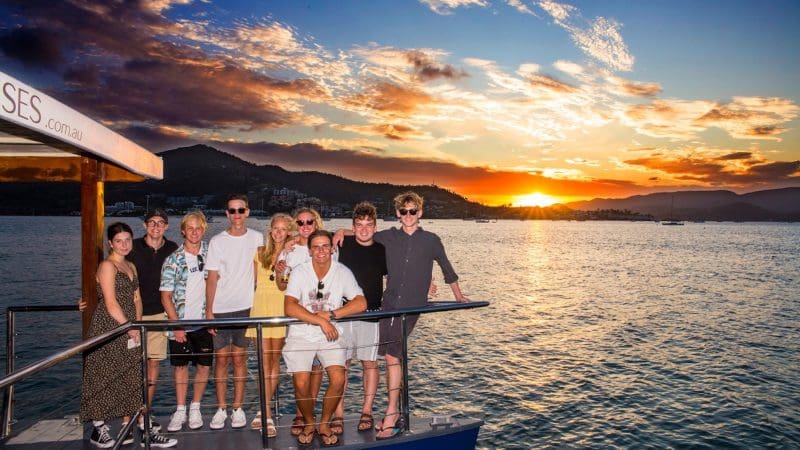 Nine friends enjoy a Sundowner Sunset Cruise, smiling as they pose with scenic mountain views and sparkling water in the background.