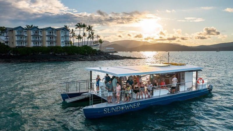 A group relishes a Sundowner Sunset Cruise along the scenic coastline, framed by palm trees and picturesque waterfront buildings.