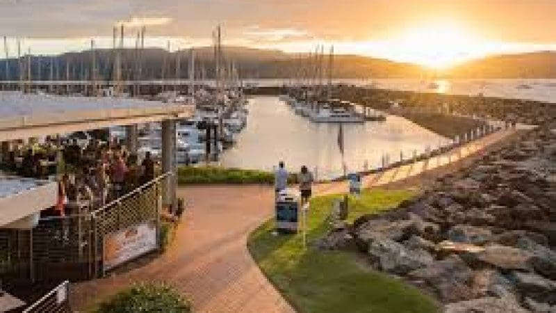 Vibrant sunset over marina with moored sailboats, people walking, and Sundowner Cruise & Dine at Sorrentos Restaurant visible on left.