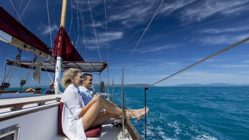 Two people unwind on Lady Enid yacht under sunny skies, overlooking turquoise waters near Whitehaven Beach after a snorkelling adventure.