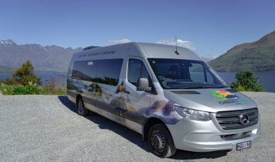 Silver tour van parked beside picturesque lake near Queenstown, snow-capped mountains, clear blue sky—scenic New Zealand travel scene.