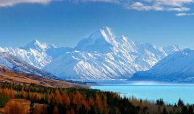 Breathtaking snow-capped mountains overlook a turquoise lake by Mt Cook, framed by vibrant autumn forests beneath a clear blue sky.