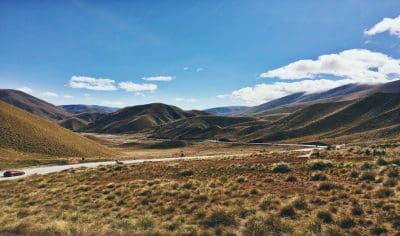 Scenic winding road through golden hills beneath clear blue skies, with a red car driving between Queenstown and Mt Cook, New Zealand.