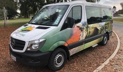 White and green nature-themed tour van parked on a picturesque, leaf-strewn roadside by Lake Tekapo with scenic views.