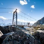 Tourists walk across a suspension bridge spanning rocky terrain, with snow-capped Mt Cook mountains and a clear blue sky overhead.