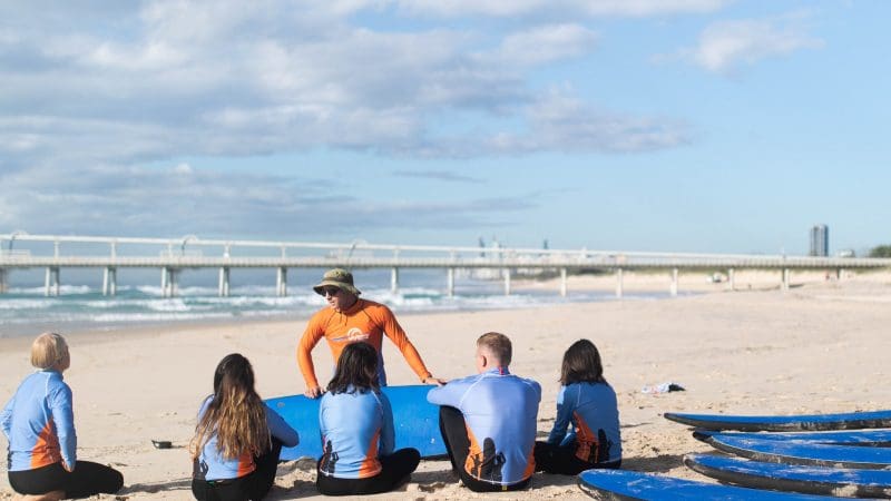 Surf instructor teaches five teenagers, aged 13 and over, during a 2-hour beginner surf lesson on a sunny beach with waves in the background.