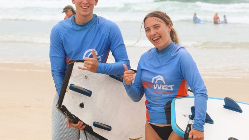 Two happy teenagers, aged 13+, smiling and giving a thumbs up after a beginner surf lesson on a sunny sandy beach, sea in view.