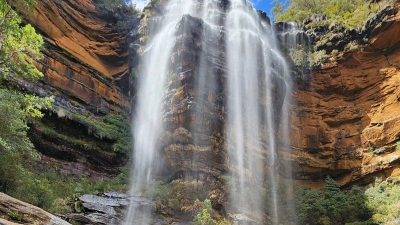 Experience a majestic, slender waterfall tumbling down rocky cliffs on a Blue Mountains Day Tour Waterfall Walk beneath a scenic sky.