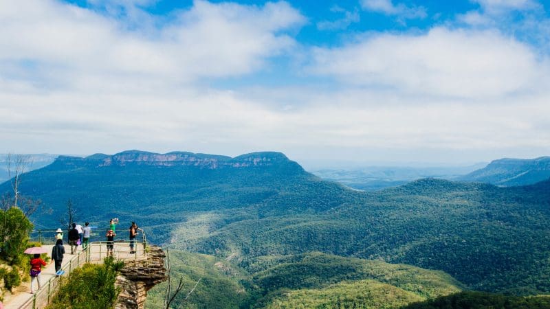 Visitors take in breathtaking views at a Blue Mountains Wildlife Park lookout, surrounded by lush green peaks and a radiant blue sky.