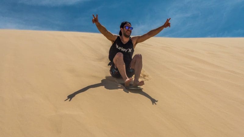 Man sandboarding down a sunlit Port Stephens dune after a 1 Day Dolphin Watching tour with Colourful Collective Travel adventure.