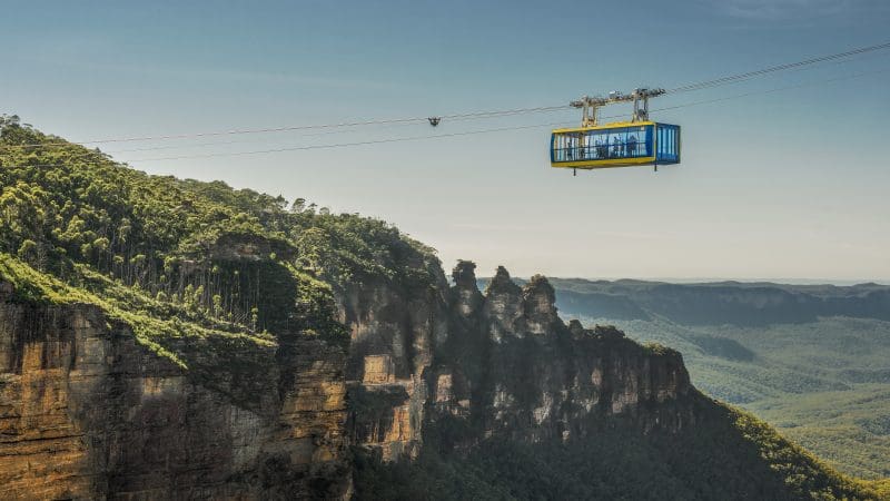 Experience breathtaking views as a vibrant blue and yellow cable car soars above Lyrebird Pass at Scenic World on a sunny day.